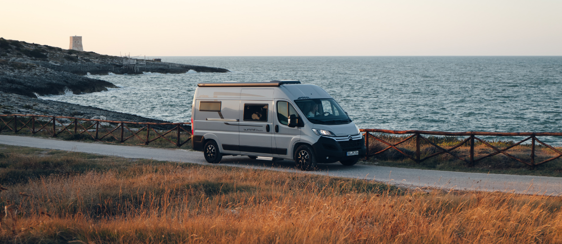 Ein grauer Campervan steht auf einer schmalen Küstenstraße, rechts davon das weite Meer mit leichtem Wellengang. Im Vordergrund wachsen trockene, goldene Gräser, im Hintergrund führt ein niedriger Holzlattenzaun die Straße entlang. Auf einer Felszunge links im Bild steht ein kleiner, heller Turm. Die Szene wirkt ruhig und warm im Licht des Sonnenuntergangs.